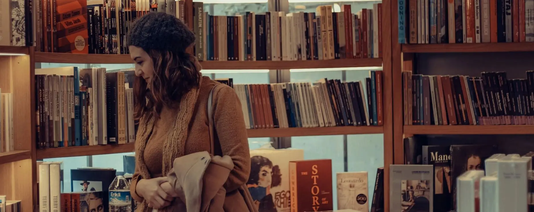 A woman is standing in a bookstore, looking at something outside the frame of the picture. In the background are bookshelves showing horizontally stacked books.