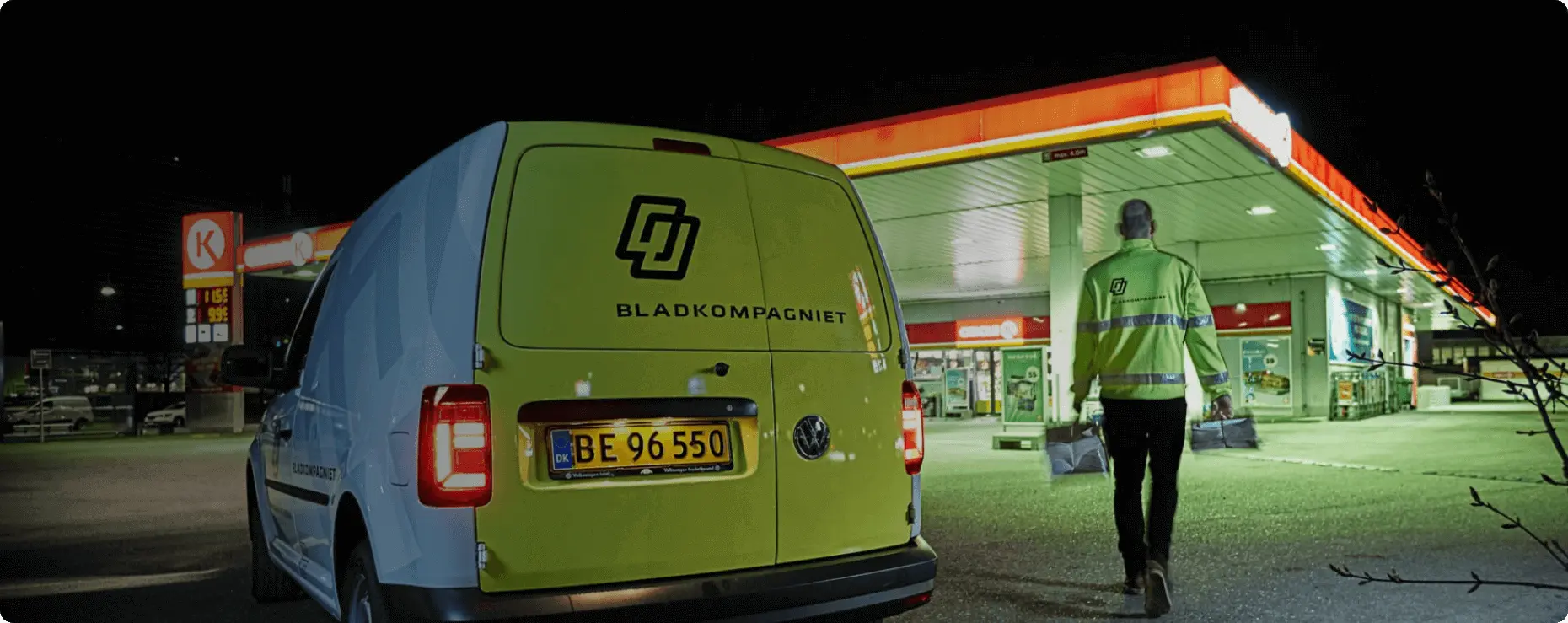 A picture of a yellow van from the back. The words Bladkompagniet are written on the van. A person in a yellow Bladkompagniet jacket is walking past the van.